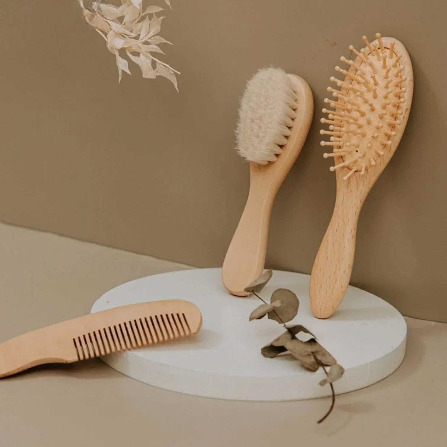 Drei hölzerne Haarwerkzeuge - ein Kamm, eine Bürste mit weichen Borsten und eine Nadelbürste - sind auf und um eine kreisförmige weiße Plattform mit getrockneten Blättern als Dekoration vor einem beigen Hintergrund angeordnet.