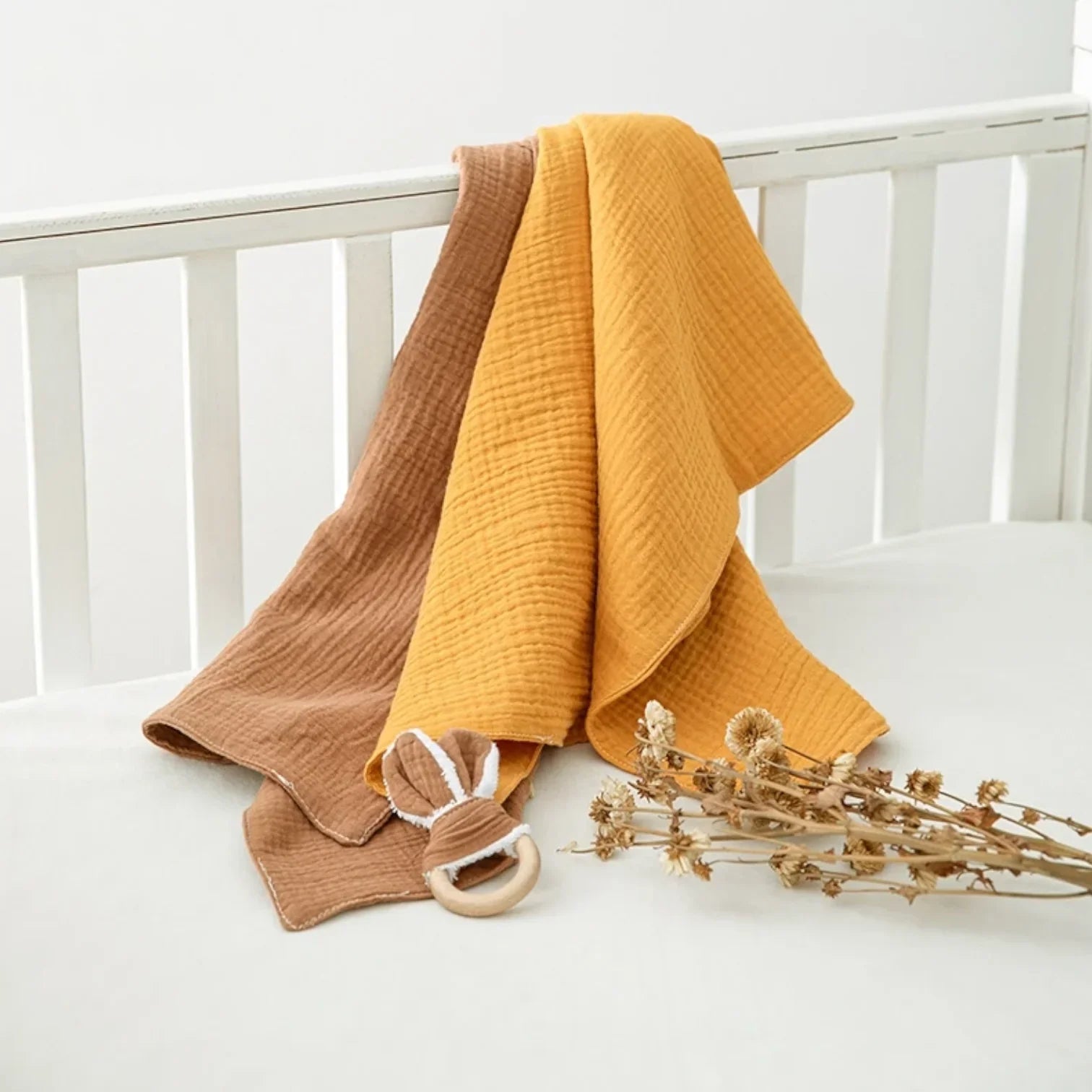 Zwei Babydecken aus Musselin, eine senfgelbe und eine braune, sind über ein weißes Krippengitter drapiert. Ein hölzerner Beißring mit Hasenohren und getrockneten Blumen liegt auf dem Laken des Kinderbetts.