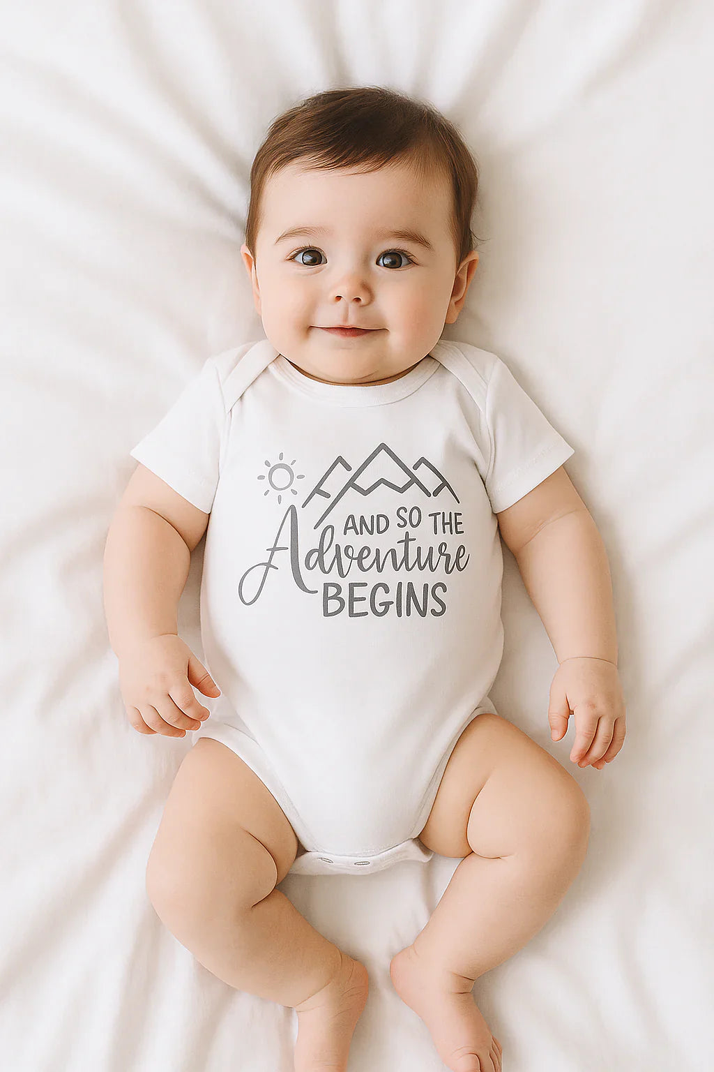 Ein lächelndes Baby liegt auf einer weißen Decke und trägt einen weißen Strampler mit einer Berggrafik und dem Aufdruck "And so the adventure begins".