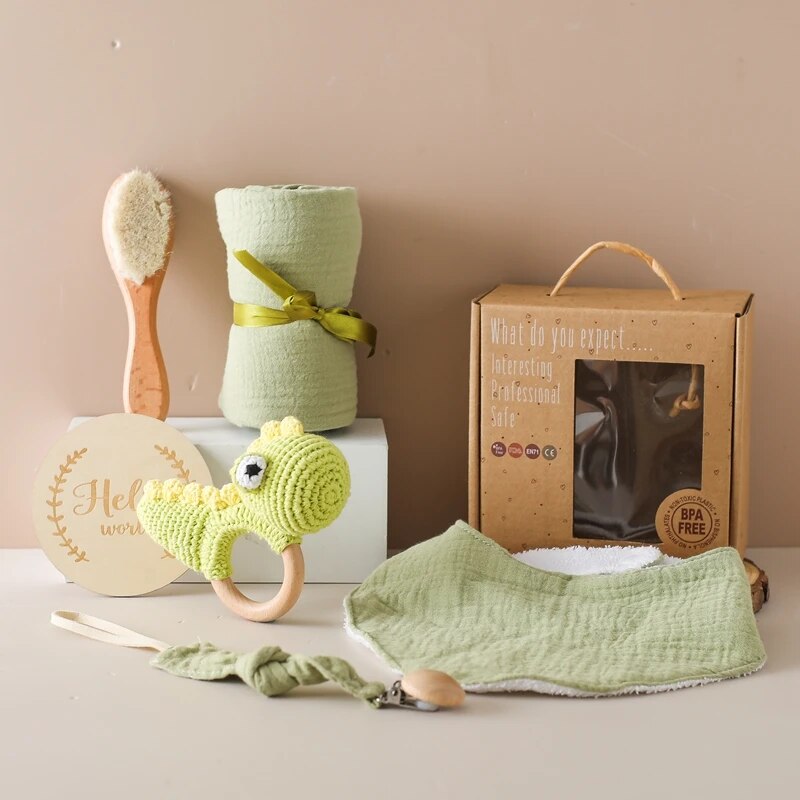 Das Hello World Alfredo Baby Geschenk zur Geburt besteht aus einer Bürste, einer grünen Decke, einer Schachtel, einem Holzschild, einer Schnullerkette, einer Dinosaurier-Rassel, einem grünen Lätzchen und einem Schnuller-Clip, alles auf einer beigen Oberfläche.