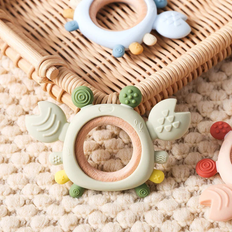 Der Beissring Krabbe Grün [11,5x8cm] von Hello World, ein weicher Beißring aus Silikon und Holz in Form einer Krabbe mit strukturierten Details, sitzt auf einem geflochtenen Korb neben anderen pastellfarbenen Beißringen.