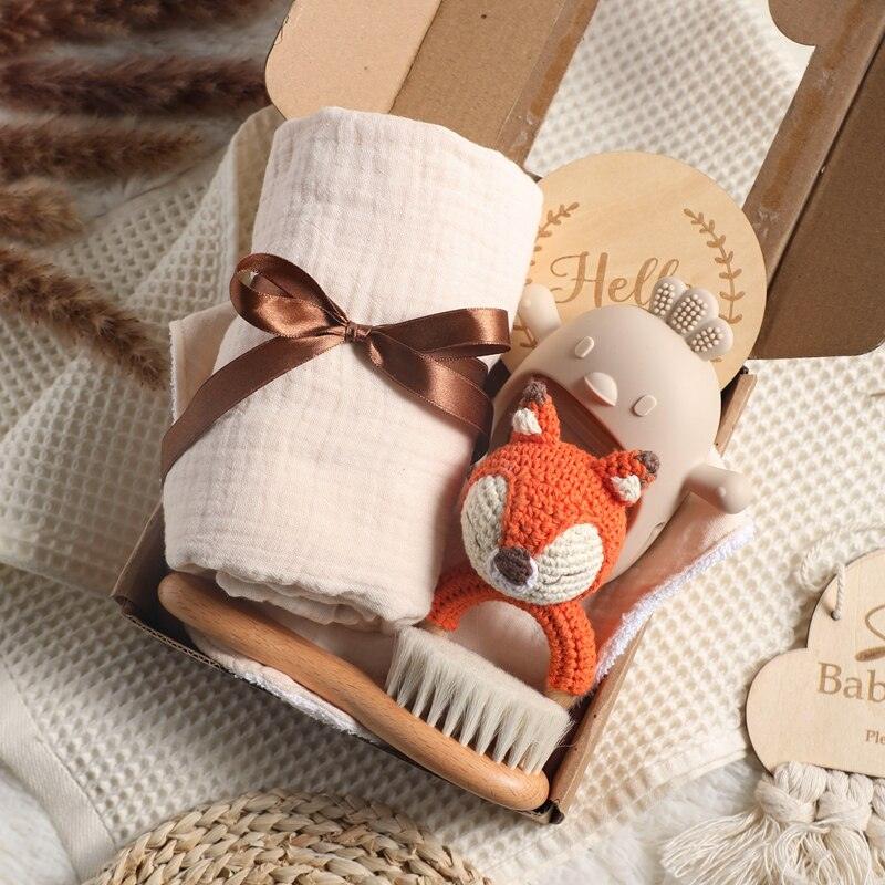 Das Foxy Baby Geschenk zur Geburt von Hello World besteht aus einem gefalteten weißen Tuch mit braunem Band, einer weichen Babybürste, einem gestrickten Rasselfuchs und einem beigen Beissring in Bärenform - perfekt zum Verschenken.