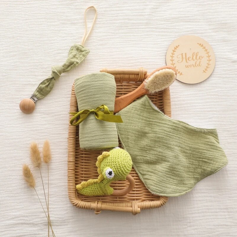 Das Alfredo Baby Geschenk zur Geburt von Hello World besteht aus einem Weidenkorb mit grünen Babyartikeln: einem Musselintuch, einer Holzbürste, einem Stofflätzchen, einem gehäkelten Dinosaurierspielzeug sowie einem "Hello World"-Holzschild und getrockneten Gräsern auf einer weißen Oberfläche.