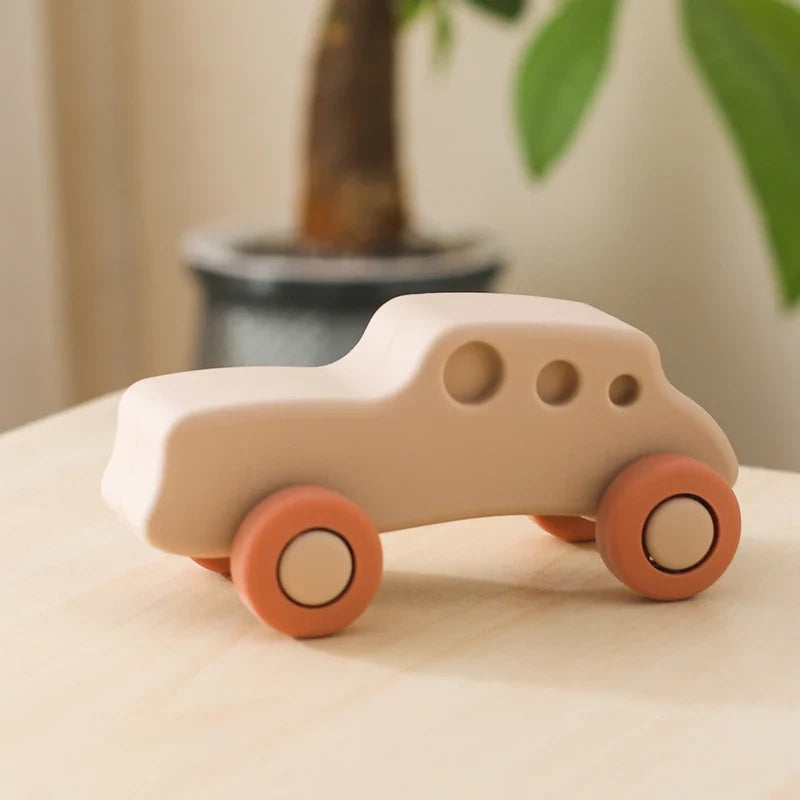 Das Auto Beige aus Silikon [11x5cm] von Hello World steht mit abgerundeten Kanten und orangen Rädern auf hellem Holz. Im Hintergrund ist eine Pflanze erkennbar. BPA-frei für sicheres Spielen Ihres Babys.