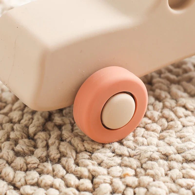 Eine Nahaufnahme des Hello World Auto Beige aus Silikon [11x5cm] mit korallenrosa Rädern, das auf einem strukturierten, gewebten beigen Teppich ruht.