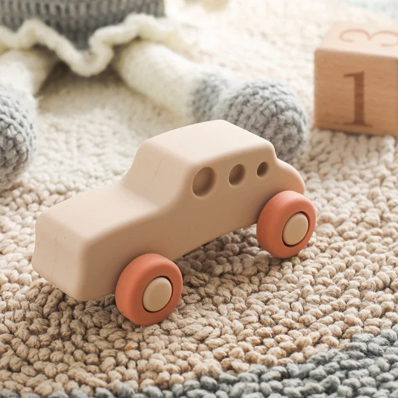 Das BPA-freie Auto Beige aus Silikon [11x5cm] von Hello World mit orangen Rädern ist das ideale Spielzeugauto zum Zahnen - perfekt neben einer Strickpuppe und einem Holzblock mit der Zahl 1 auf dem Teppich.
