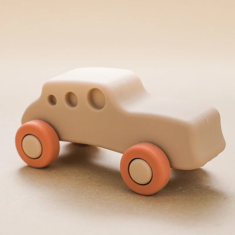 Das Hello World Auto Beige aus Silikon [11x5cm] hat ein minimalistisches beiges Design mit runden orangefarbenen Rädern und drei kreisförmigen Fenstern - ideal als Spielzeugauto Baby oder Beißspielzeug, dargestellt auf einer hellbeigen Oberfläche.