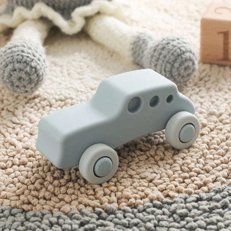 Ein weiches, hellblaues Auto Blau aus Silikon [11x5cm] von Hello World mit runden Rädern steht auf einem strukturierten beige- und hellbraunen Teppich, mit gestrickten Puppenbeinen und einem Holzklotz im Hintergrund.
