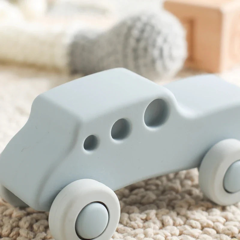 Ein hellblaues Auto Blau aus Silikon [11x5cm] von Hello World sitzt auf einem beigen Teppich mit verschwommenen Plüschtieren und einem Holzklotz - perfekt als Spielzeugauto Baby oder sanfte Beißhilfe.