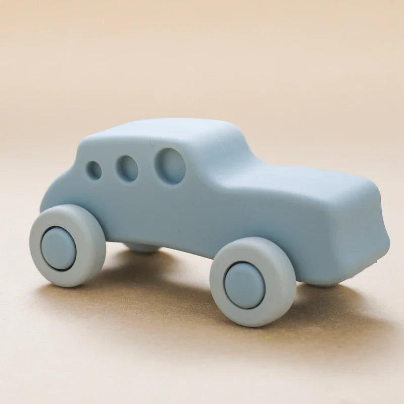 Ein hellblaues Auto Blau aus Silikon [11x5cm] von Hello World mit runden schwarzen Rädern erscheint auf einem neutralen beigen Hintergrund. Das minimalistische Spielzeugauto hat einfache, abgerundete Formen und drei Fenster - ideal für ein Spielzeugauto Baby.