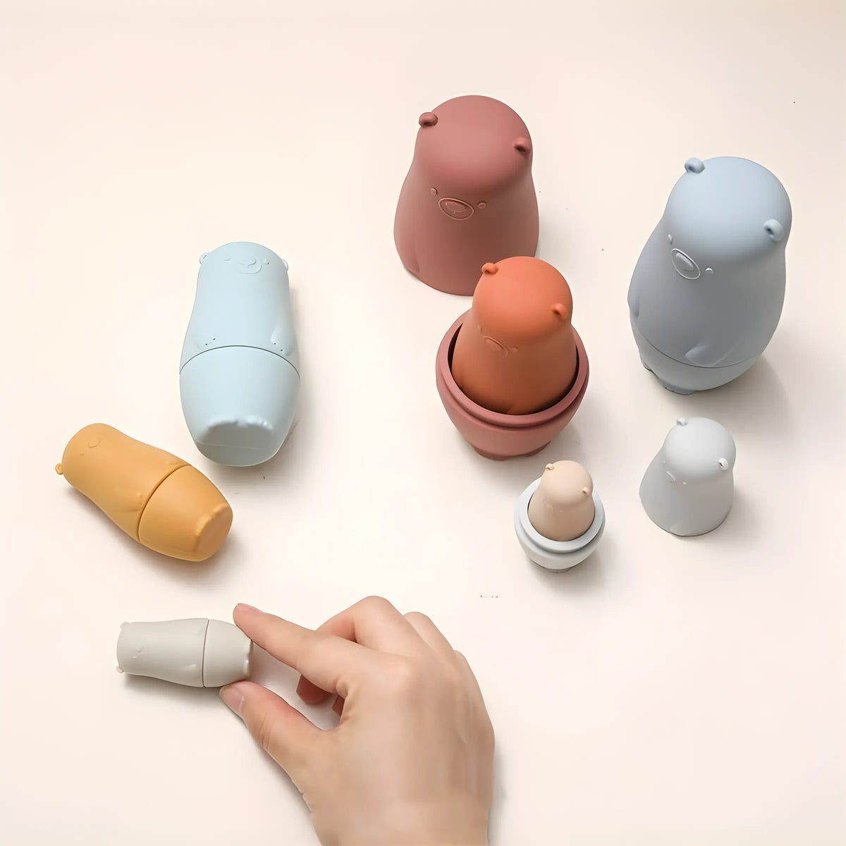 Die Hello World Bär Rot aus Silikon [6x13cm] Nesting-Figuren in sanften Farbtönen werden auf einer hellen Oberfläche angeordnet; einige sind gestapelt, andere stehen auseinander und zeigen ihr Babuschka-artiges Design - ideal als entzückendes Lernspielzeug.