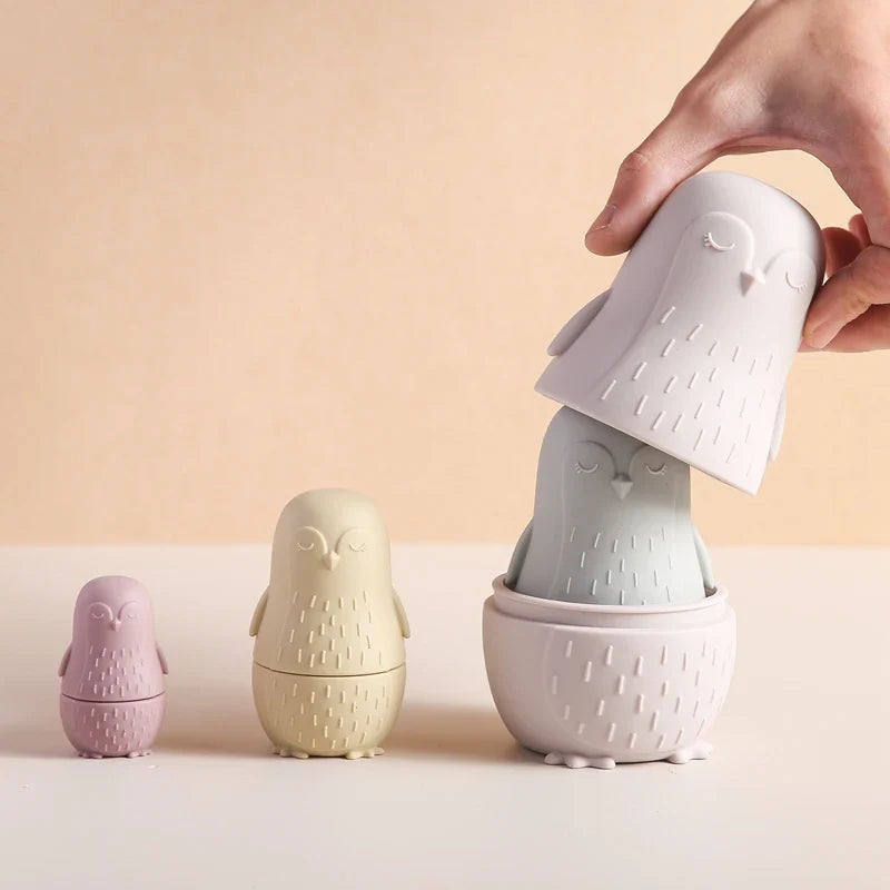 Eine Hand hebt die obere Hälfte des größten Hello World Pinguin Beige aus Silikon [7x12cm] an und enthüllt einen kleineren Pinguin im Inneren; drei pastellfarbene Pinguine sind in einer Reihe vor einem weichen beigen Hintergrund angeordnet.