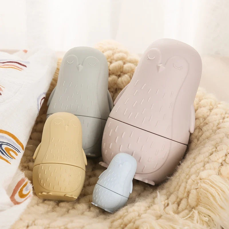 Der Hello World Pinguin Beige aus Silikon [7x12cm] sitzt auf einer weichen, cremefarbenen Strickdecke, neben der ein gemusterter Stoff hervorschaut.