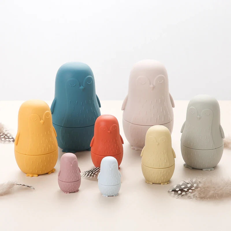 Der "Pinguin Beige aus Silikon [7x12cm]" von Hello World wird auf einer hellen Oberfläche präsentiert. Er besticht durch seinen sanften Pastellton und sein charmantes Design, das von verstreuten Federn umgeben ist, die ihm einen skurrilen Touch verleihen.