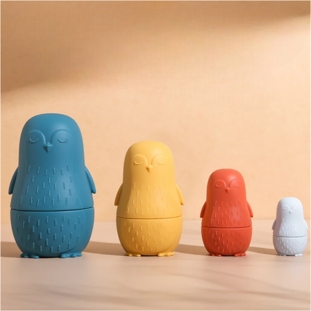 Der Hello World Pinguin Blau aus Silikon [7x12cm] sitzt zwischen blauen, gelben, roten und weißen Babushka-Pinguin-Puppen, die nach Größe sortiert auf einem neutralen Hintergrund mit weichen Schatten angeordnet sind - für tollen Stapel- und Sortierspaß.