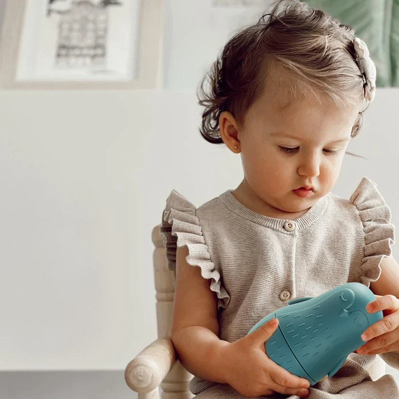 Ein kleines Kind mit dunklen Haaren sitzt auf einem Stuhl, trägt ein beiges Outfit mit gerüschten Ärmeln und blickt auf das Spielzeug Hello World Pinguin Blau aus Silikon [7x12cm] in seinen Händen.