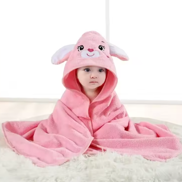 Ein Baby sitzt auf einer weichen Decke, eingewickelt in das rosafarbene Badetuch mit Kapuze Hase [80x80cm] von Hello World, das ein süßes Hasengesicht, Ohren und gestickte Details aufweist. Sanftes Licht füllt den Hintergrund, während das Baby mit neutralem Blick nach vorne schaut.