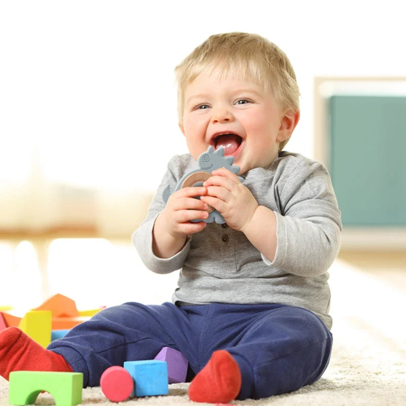 Ein lächelndes blondes Kleinkind sitzt auf einem Teppich und kaut auf dem Hello World Beissring Dino Blau [9x9cm]. Bunte Holzklötze liegen in der Nähe, und das Kind trägt ein graues Hemd, eine blaue Hose und rote Socken.