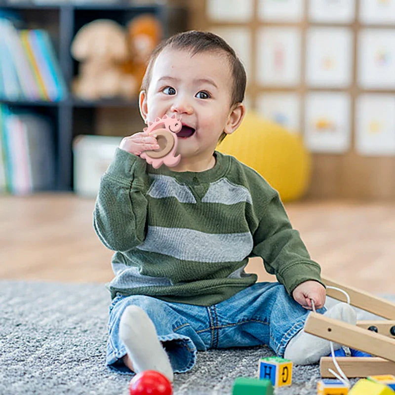 Ein Baby in einem grün-grau gestreiften Hemd und blauen Jeans sitzt auf einem Teppich und hält lächelnd den Hello World Beissring Dino Rosa [9x9cm] in der Hand. Im Hintergrund sind bunte Spielzeuge und ein Bücherregal zu sehen.
