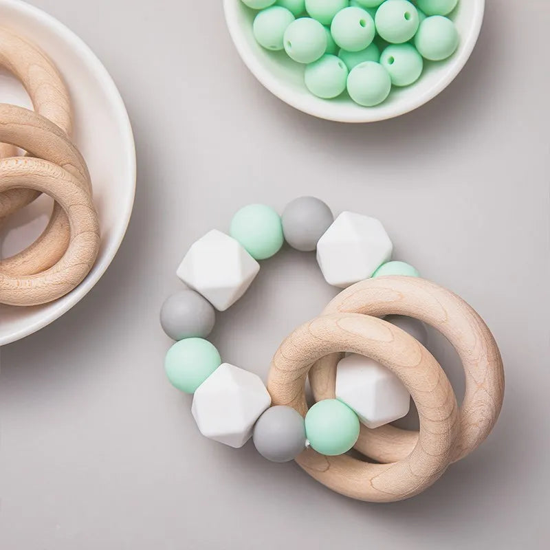 Der Hello World Beissring Kette Duo Mintgrün [7x9cm] ist ein mintgrüner, weißer und grauer Beißring aus Silikon mit Holzringen und Perlen, der auf einem hellen Hintergrund neben Schalen mit zusätzlichen Perlen und Holzringen abgebildet ist.