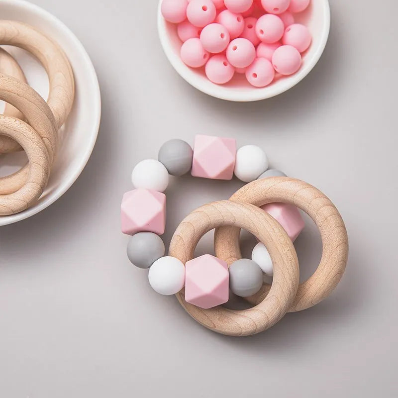 Die Hello World Beissring Kette Duo Rosa [7x9cm] mit rosa, grauen und weißen Perlen ist eine bezaubernde Beißhilfe auf hellem Grund neben einer Schale mit runden rosa Perlen.