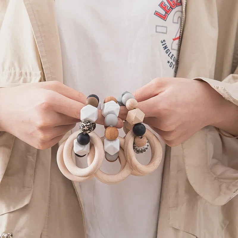Eine Person in beiger Jacke hält drei Hello World Beissring Kette Duo Schwarz (7x9cm), jeweils mit Holz- und Silikonperlen in neutralen Farben - ideal als Zahnungshilfe Baby oder Beißring-Kette.