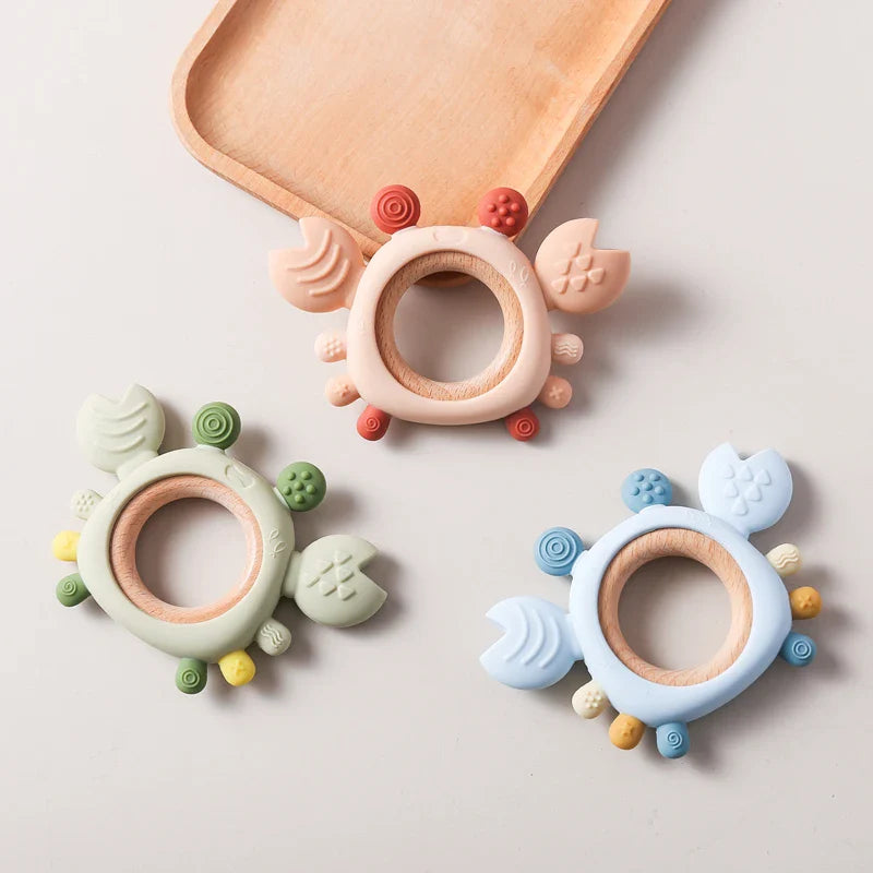 Der Hello World Beissring Krabbe Beige (11,5 x 8 cm) ist ein Beißspielzeug im beigen Krabbendesign mit Holz- und Silikonelementen, das auf einer hellen Fläche neben einem Holztablett angeordnet ist.