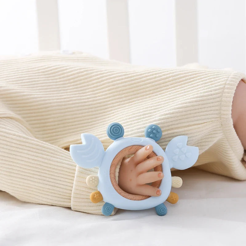 Ein Baby in einem cremefarbenen, gerippten Outfit hält den Hello World Beissring Krabbe Blau [11,5x8cm], einen krabbenförmigen Beißring aus Silikon in Hellblau und Beige mit strukturierten Enden, der perfekt in die kleine Hand des Babys passt.