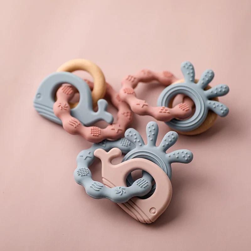 Drei pastellfarbene Baby-Beißringe aus Silikon, darunter der Hello World Beissring Walfisch Blau (10,5x7cm), sind auf einer rosa Oberfläche angeordnet. Sie haben eine strukturierte Oberfläche und runde Griffe in Hellblau und Blassrosa.