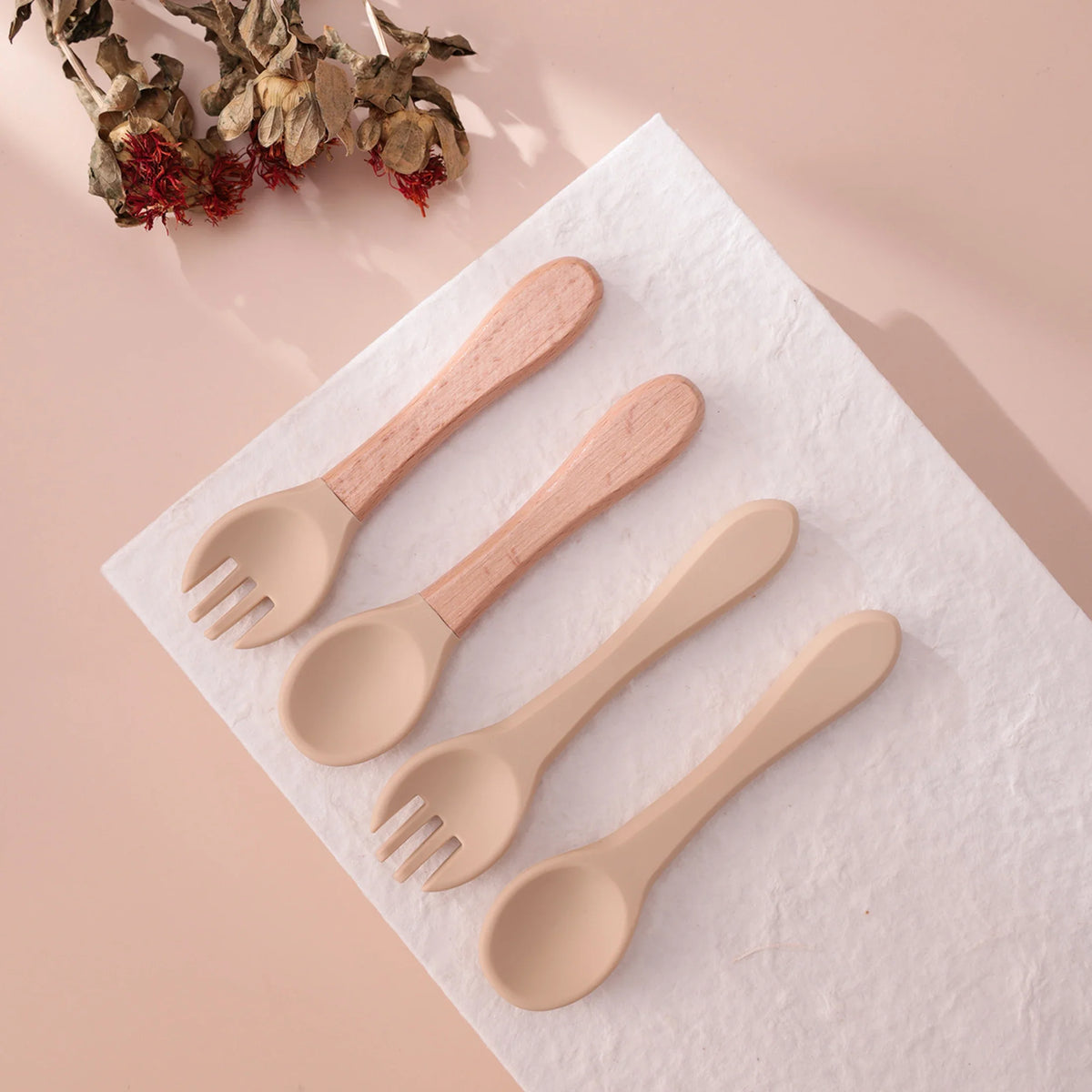 Vier Hello World Besteck Beige Silikon [3,5x14cm] Löffel mit Holzgriffen, alle BPA-frei, sind auf einem weißen strukturierten Tuch mit getrockneten Blumen auf einem blassrosa Hintergrund angeordnet. Perfekt als Baby Besteckset.