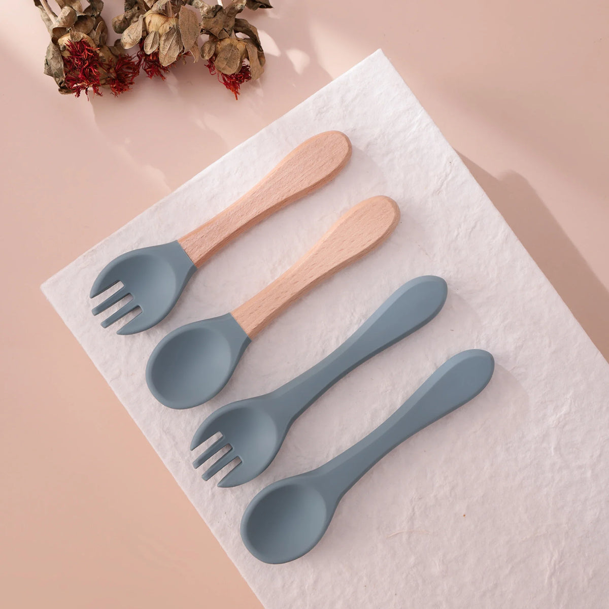 Zwei Sets Hello World Besteck Blau Silikon [3,5 x 14 cm] - eine Gabel und ein Löffel mit Holzgriffen - sind auf einer weißen, strukturierten Oberfläche ausgestellt, ergänzt durch getrocknete Blumen in der oberen linken Ecke.