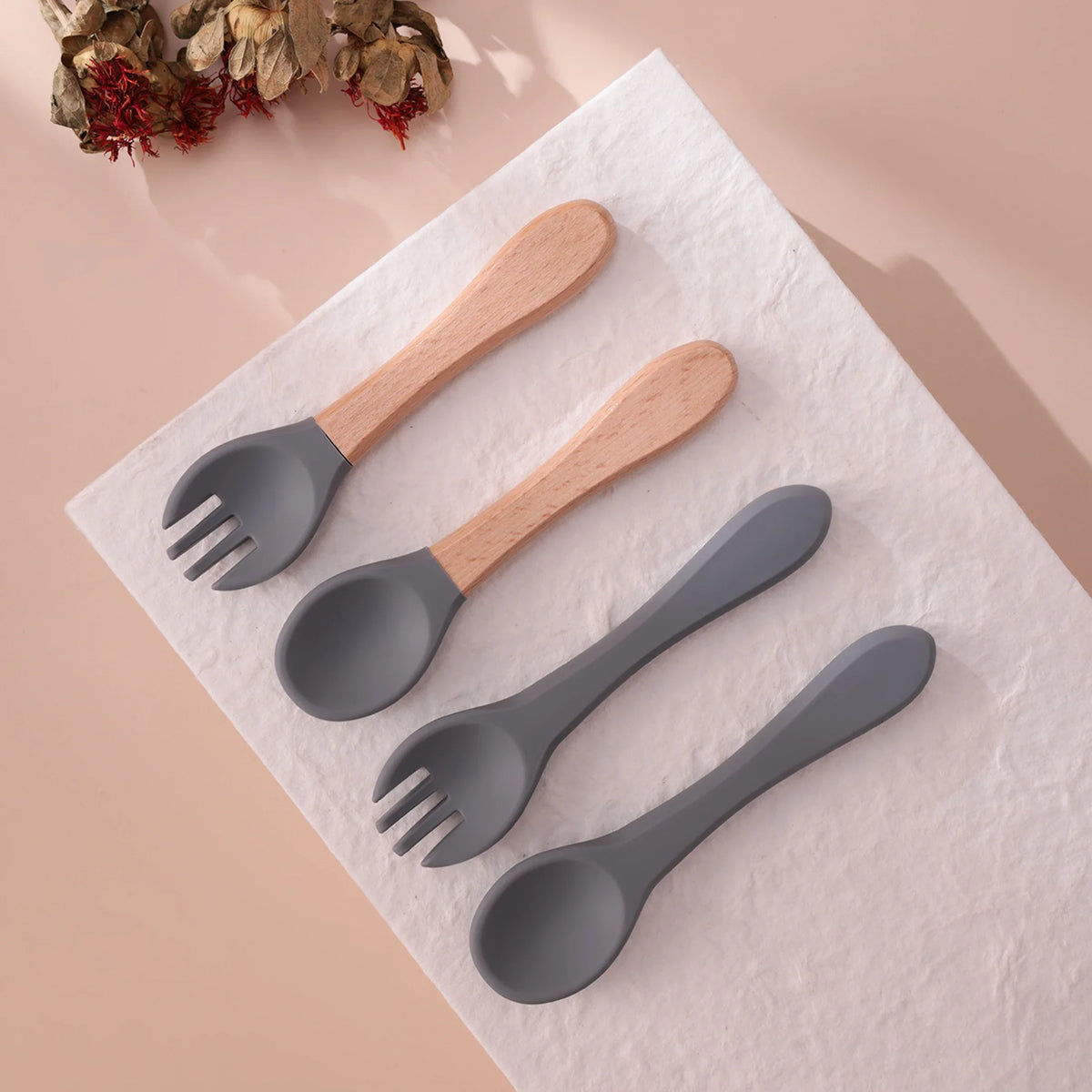 Vier graue "Hello World" Bestecke aus Holz und Silikon [3,5x14cm] bilden ein stilvolles, nachhaltiges Set auf weißem Untergrund. Im oberen linken Eck liegen getrocknete Blumen auf beigem Grund.