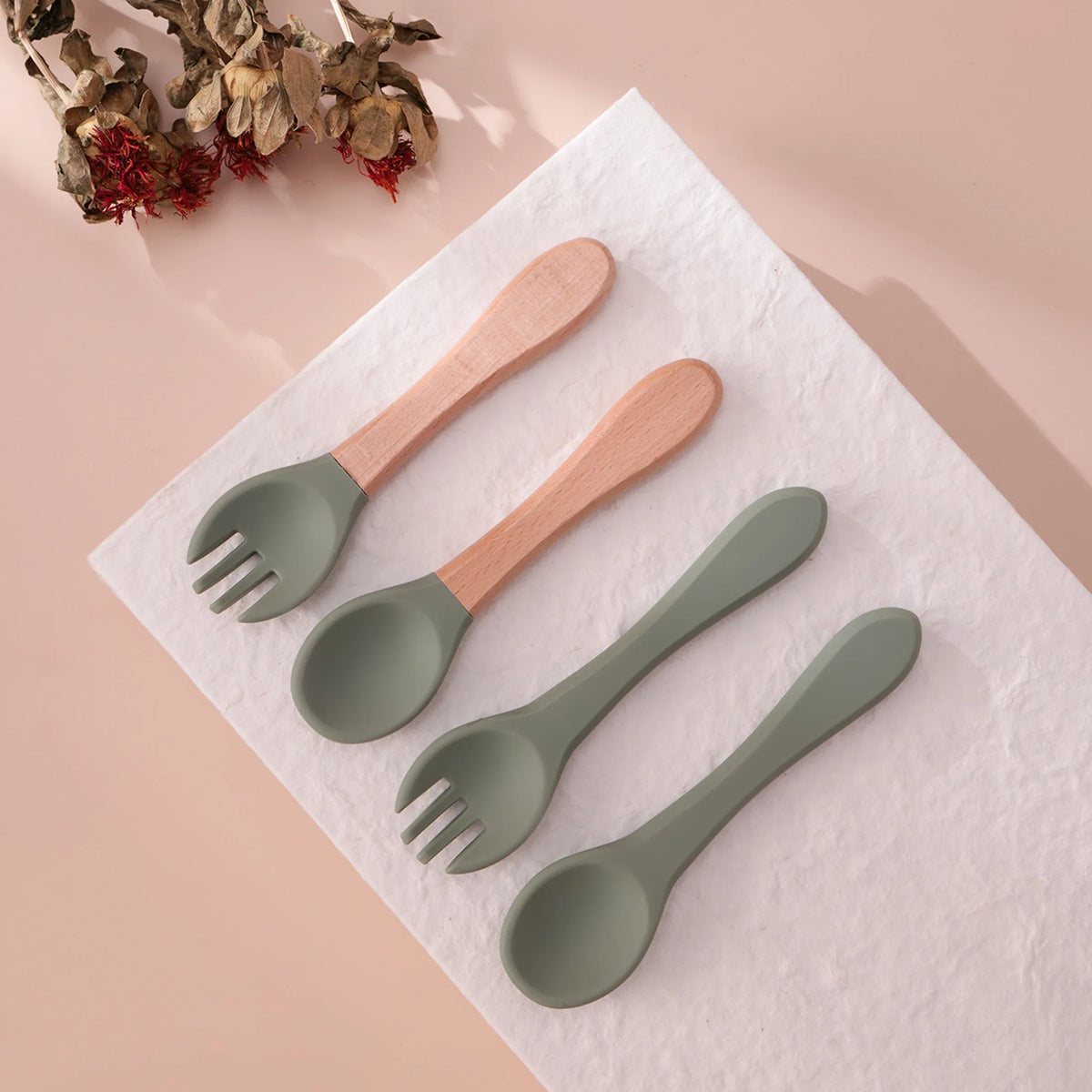 Das umweltfreundliche Kinderbesteck-Set "Besteck Grün Holz/Silikon [3.5x14cm]" von Hello World wird ordentlich mit Trockenblumen auf weißer Fläche präsentiert - zwei Holzgriffe, ein grünes Besteck, perfekt für kleine Esser.