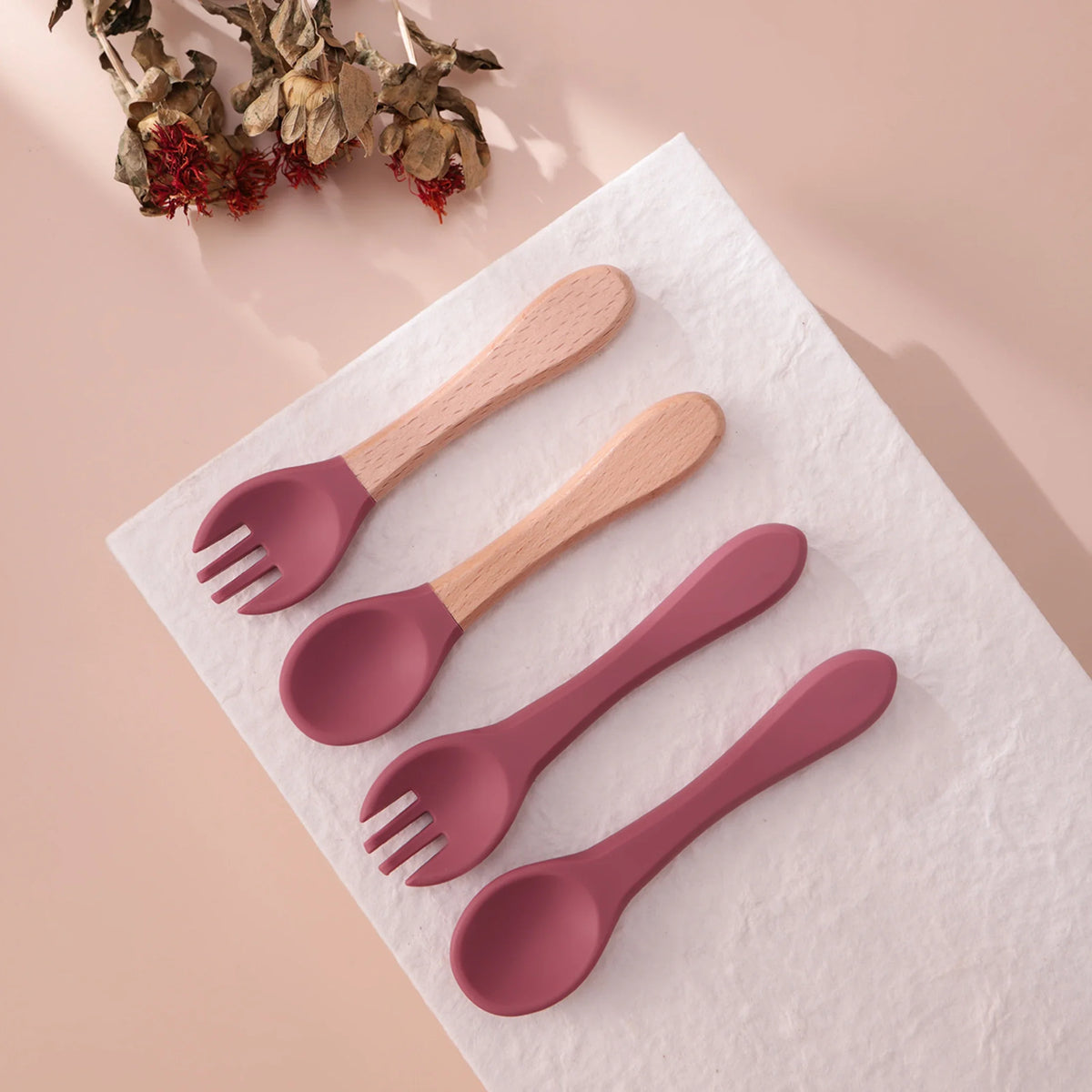 Drei Sets Hello World Besteck Pink Silikon [3,5 x 14 cm] - zwei mit Holzgriffen, eines in unifarbenem Rosa - sind ordentlich auf einer weißen, strukturierten Oberfläche mit getrockneten Blumen arrangiert und bilden ein bezauberndes Baby Besteck Display.