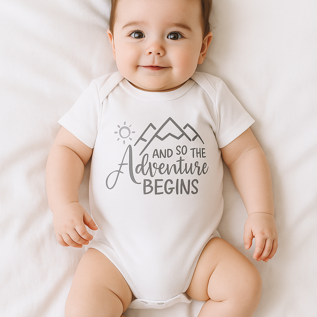 Ein lächelndes Baby liegt auf einer weißen Decke und trägt den Hello World Body [33,5x38cm] (6-9 Monate) aus weicher Baumwolle mit Druckknöpfen und Mountain-and-Sun-Print: "Und so beginnt das Abenteuer.