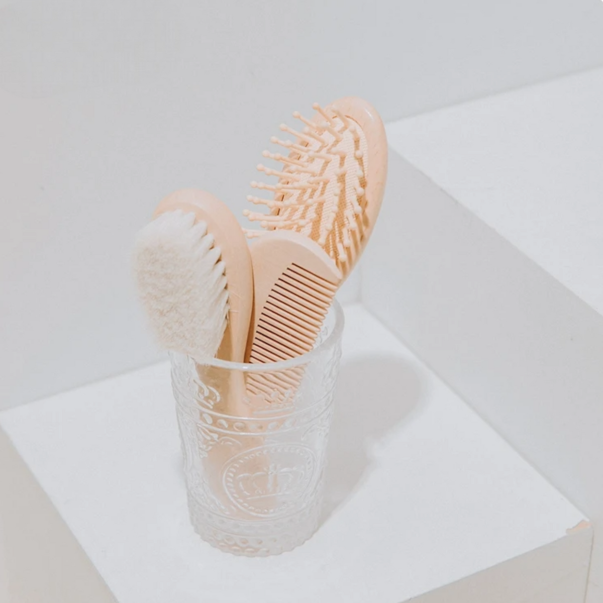 In einem durchsichtigen Glas befinden sich die Hello World Haarbürste - Kugelborsten [5x18cm], eine Bürste mit weichen Borsten und ein Kamm in hellen Beigetönen, die alle ordentlich vor einem minimalistischen weißen Hintergrund angeordnet sind.