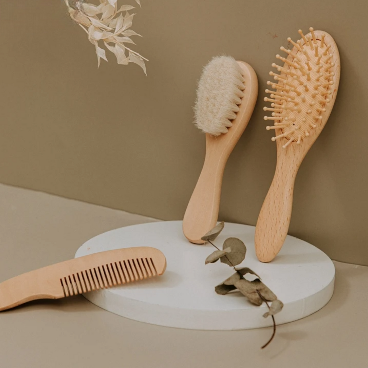 Drei Hello World Haarbürsten - Kugelborsten [5x18cm] aus Holz werden auf einem runden weißen Podest mit getrockneten Blättern ausgestellt, wodurch ein minimaler, natürlicher Look vor einem beigen Hintergrund entsteht.