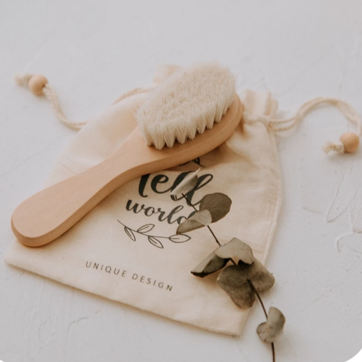 Die Hello World Haarbürste - Weichborsten [4x15cm] liegt auf einem beigen Stoffbeutel mit Aufdruck und Blattdesign, umgeben von Eukalyptusblättern - ideal für sanfte Pflege auf hellem Untergrund.