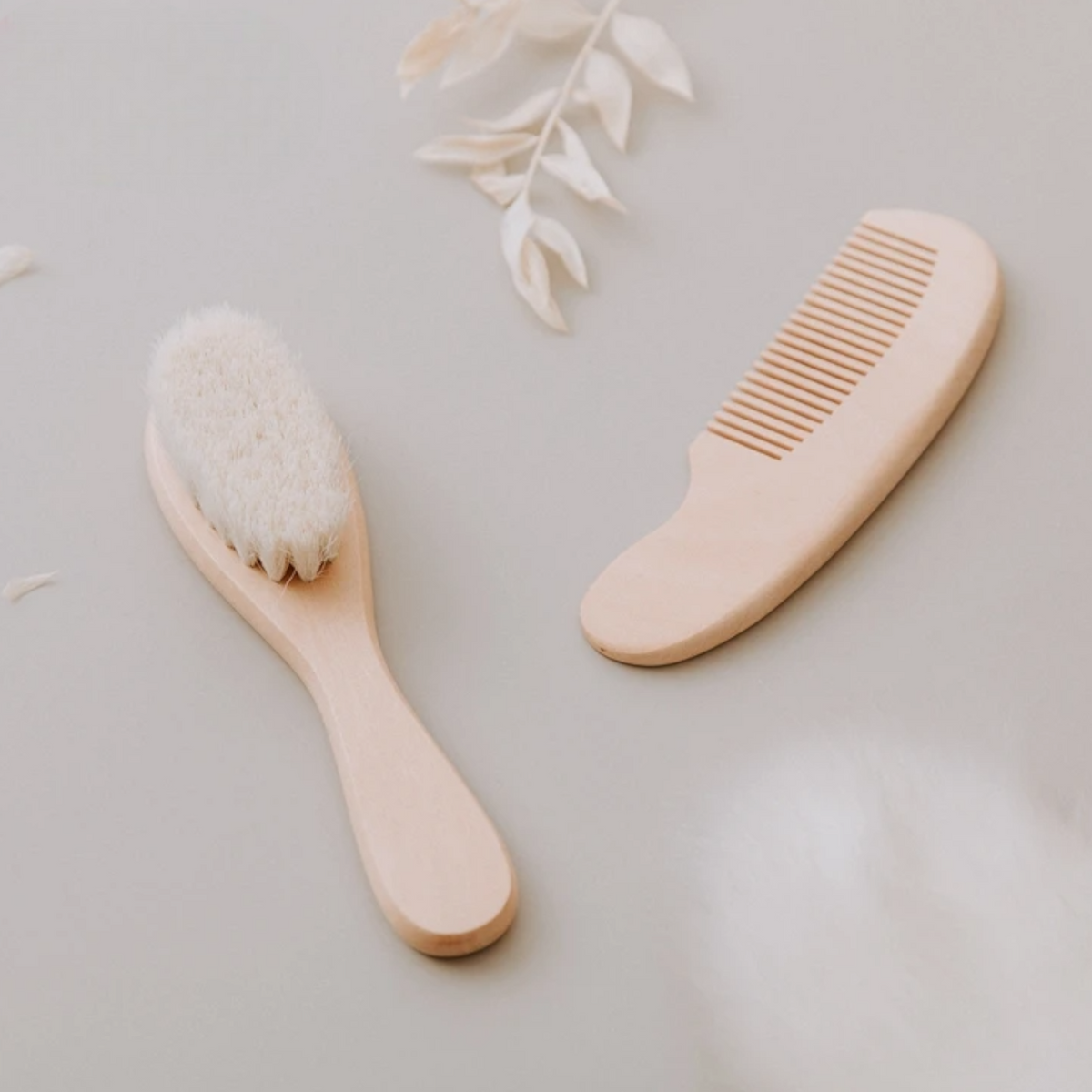 Die Hello World Haarbürste - Weichborsten [4x15cm] mit Holzkamm liegt auf heller Fläche, umgeben von weißen Zierblättern und flauschigen Akzenten - ideal zur sanften Pflege empfindlicher Babyhaare.