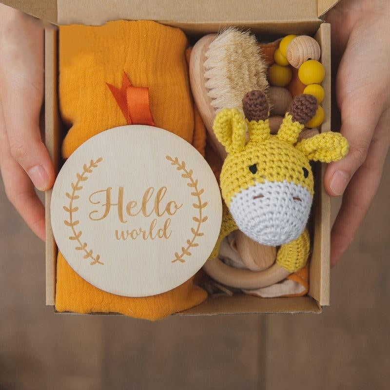 Ein Paar Hände hält das Carl Baby Geschenk zur Geburt von Hello World: eine Geschenkbox mit einer gelben Babydecke, einem Beißring aus Silikonholz, einer Holzbürste, einer gelben Giraffenrassel und einer runden "Hello World"-Plakette mit Blattmotiv.