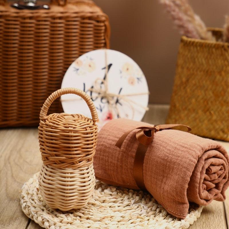 Auf einer Holzfläche unterstützt das Hello World Carla Baby Geschenk zur Geburt Set mit einem kleinen geflochtenen Korb, rostfarbenem Stoff und Holzrassel die sensorische Entwicklung. Körbchen und ein Blumenteller bilden einen charmanten Hintergrund.