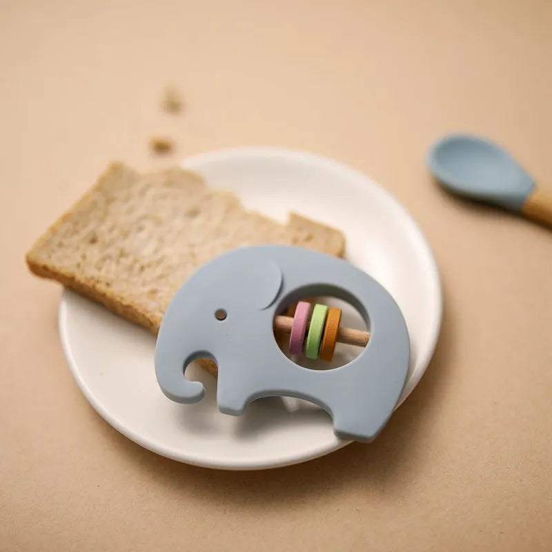 Eine Scheibe Brot auf einem weißen Teller liegt neben einem Hello World Elefant Blau aus Silikon [9x6,8cm] mit bunten Ringen in der Mitte und einem kleinen Löffel, die alle auf einer beigen Fläche angeordnet sind.