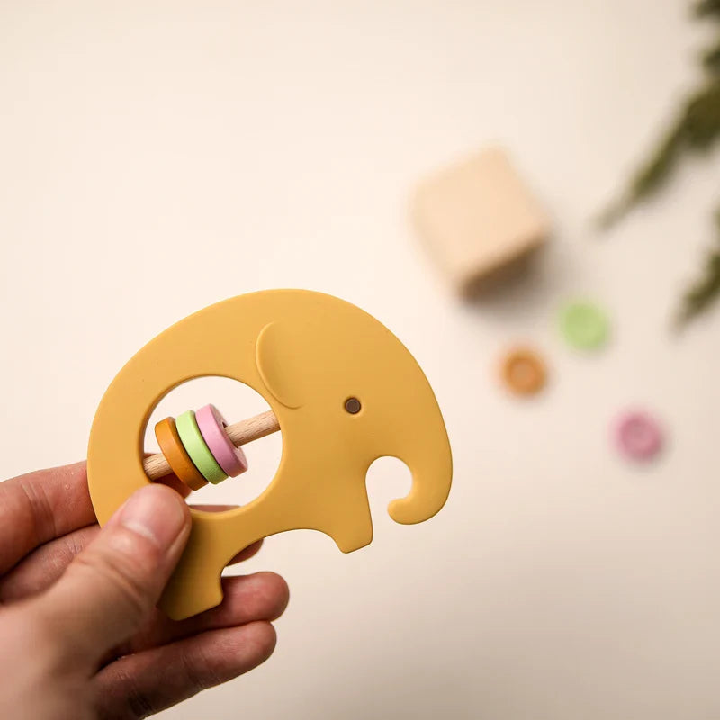 Eine Hand hält die Hello World Elefant Gelb aus Silikon [9x6,8cm] Babyrassel mit pastellfarbenen Ringen. Im unscharfen Hintergrund ruhen Babyspielzeug wie Holzklötze, rundes Spielzeug und eine grüne Pflanze auf einer hellen Fläche.