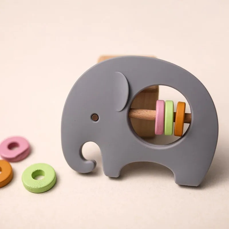Der Hello World Elefant Grau aus Silikon [9x6,8cm] hält eine Holzstange mit pinken, gelben und grünen Ringen; weitere bunte Ringe liegen auf der hellen Oberfläche bereit.