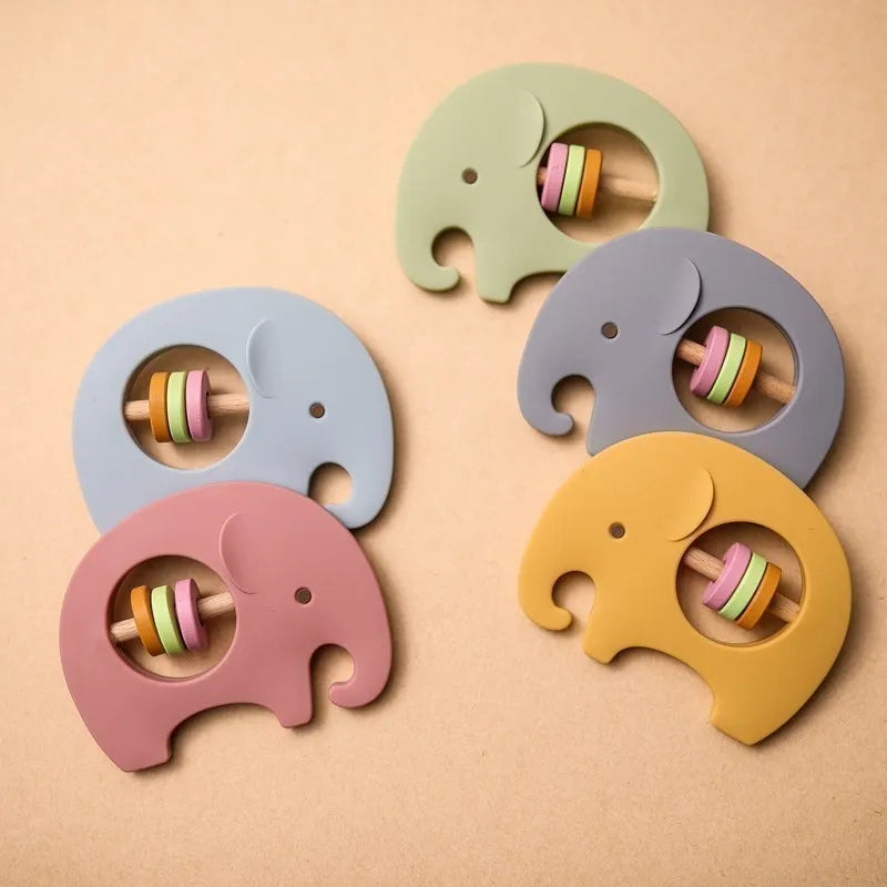 Vier "Hello World" Elefant Grau aus Silikon Rasseln (9x6,8cm) in Pastelltönen sind auf einer beigen Oberfläche angeordnet. Jede verfügt über eine kreisförmige Aussparung mit bunten Ringen im Inneren für sensorisches Spiel.