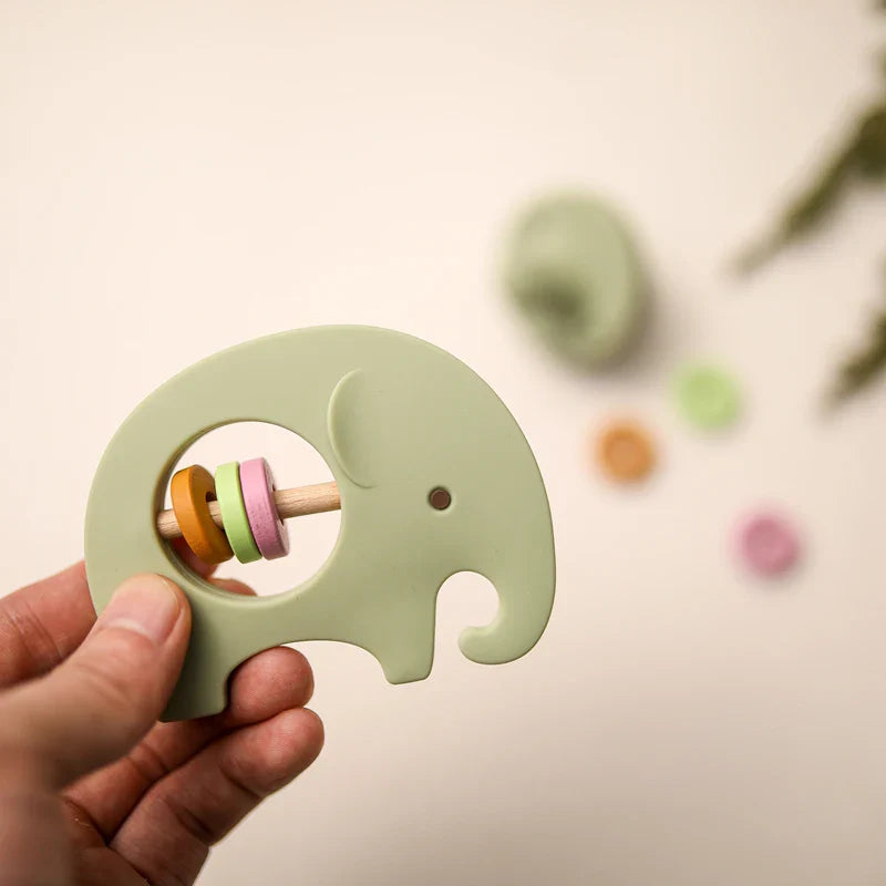 Eine Hand hält die Babyrassel Hello World Elefant Grün aus Silikon [9x6,8cm] mit pastellfarbenen Ringen; im unscharfen Hintergrund sind bunte Ringe und ein weiterer grüner Elefant Beißring auf einer hellen Fläche zu sehen.