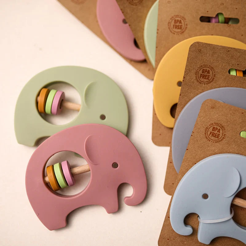 Der Baby-Beißring Hello World Elefant Grün aus Silikon [9x6,8cm] besteht aus einem Holzstab mit Ringen und ist auf braunem Karton mit BPA-FREI-Stempel verpackt, der auf einer hellen Oberfläche ausgestellt ist.
