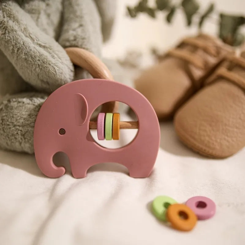 Der Hello World Elefant Rosa aus Silikon [9x6,8cm] mit bunten Ringen ruht auf einer weichen Unterlage neben einem Plüschtier und hellbraunen Babyschuhen - das perfekte Geschenk für Kinder.