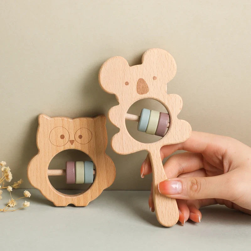 Zwei Babyrasseln aus Holz, darunter die Hello World Eule aus Holz (8x8cm) und ein Koala mit Griff, beide mit pastellfarbenen Perlen im Inneren. Eine Hand hält die Koala-Rassel auf einem beigen Hintergrund - ideales Naturholz Spielzeug.
