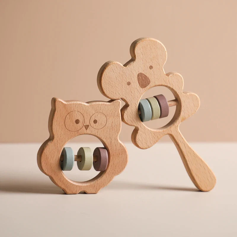 Zwei nachhaltige Baby-Rasseln von Hello World, eine Eule aus Holz [8x8cm] und ein Koala, beide mit runden Ausschnitten und pastellfarbenen Perlen vor sanft beige beleuchtetem Hintergrund.